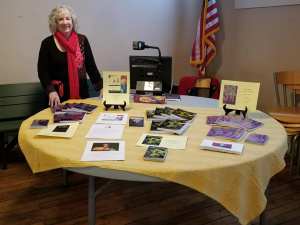 Lynda Lambert stands beside her book display on the table at a book siging event. She is wearing a red wool jacket made in Austria. She is smiling and her books are arrayed on a round table covered with a yellow cloth. 